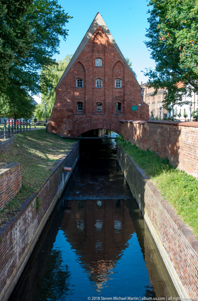 Large steep-roofed brick mill building spanning a narrow canal, with its gable reflected in the water.
