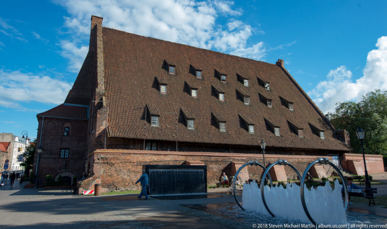 Large historic brick mill with a vast steep tiled roof and many small roof openings beside a pedestrian area.