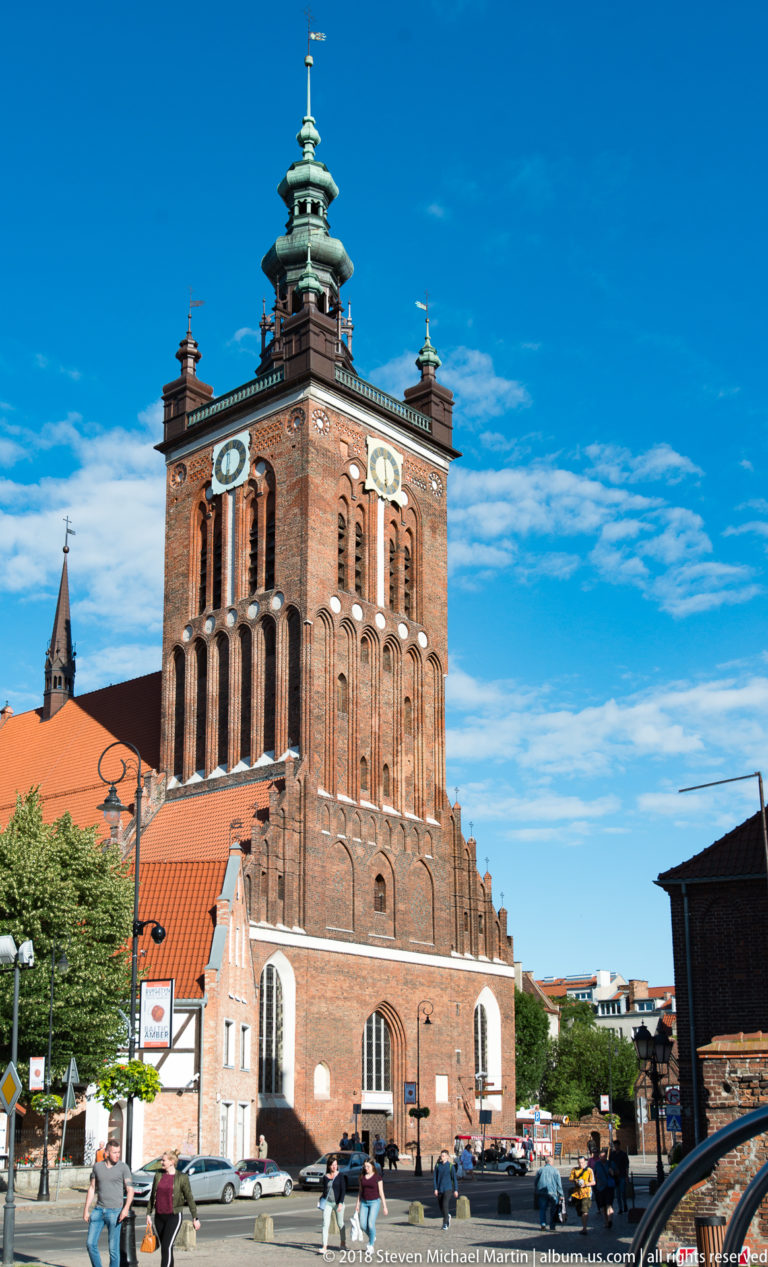 Massive square brick church tower with clock faces, arched openings, red roofs, and a green copper lantern.