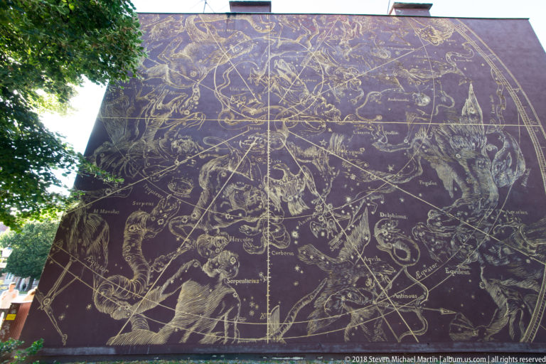 Large exterior wall mural resembling a historical star chart, filled with constellations, labels, and celestial figures.