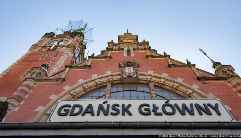 Low-angle view of Gdańsk Główny railway station entrance with large name sign and ornate brick tower.