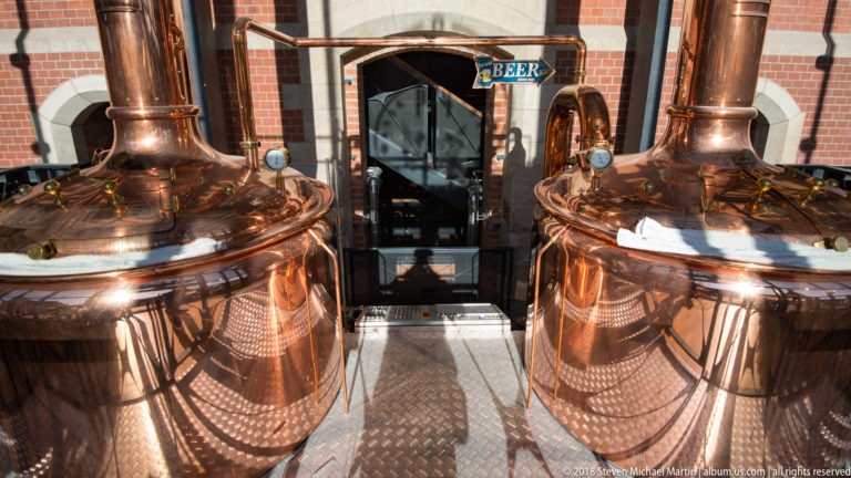 Two large polished copper brewing kettles with gauges and fittings inside a brewery, with a BEER sign between them.