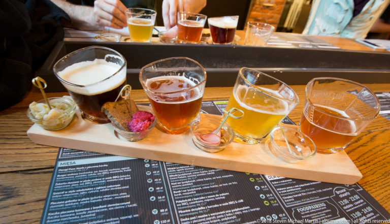 Wooden beer flight with several small glasses ranging from pale gold to dark brown, set above a printed menu.