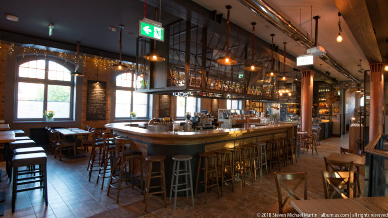 Long brewery bar with stools, pendant lights, exposed brick, metal framing, bottles, and visible brewing equipment.