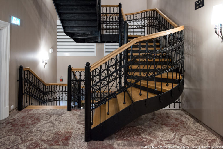 Curving wooden staircase with black cast-iron railings, multiple landings, pale walls, and patterned carpet.