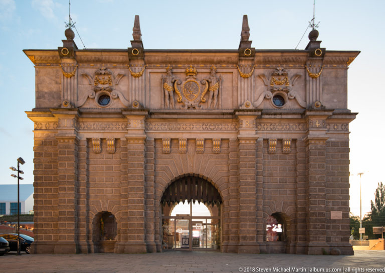 Upland Gate with rusticated stone façade, large central arch, two smaller openings, heraldic reliefs, and pointed finials.