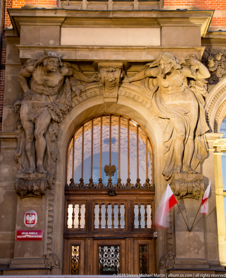 Baroque stone doorway with muscular carved figures, draped ornament, dark wooden doors, and Polish flags.