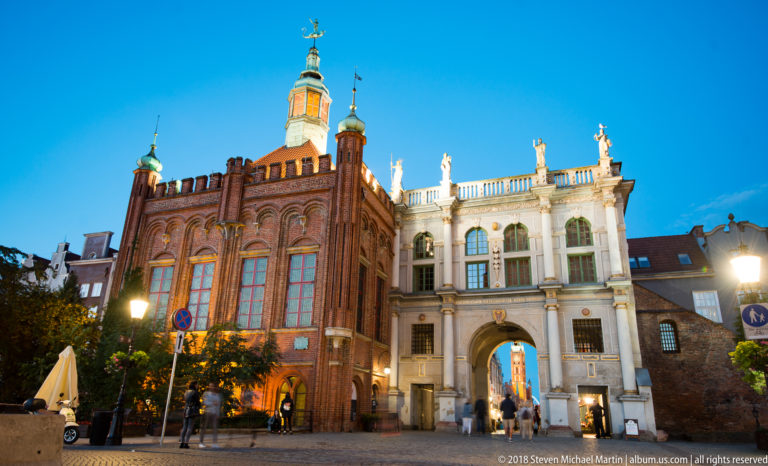 Golden Gate beside a tall red-brick Gothic building with pinnacles and tower, illuminated in evening light.
