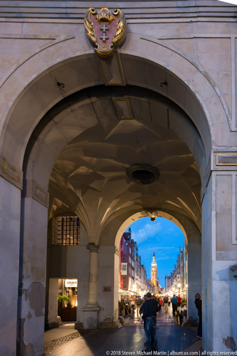 View through the Golden Gate arch toward illuminated Długa Street, with pedestrians and a distant tower at dusk.