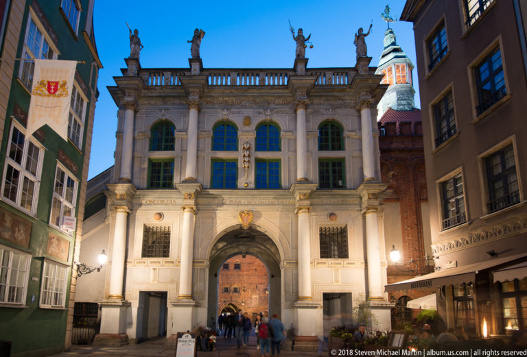 Illuminated pale stone Golden Gate with central arch, columns, rooftop statues, and historic buildings on both sides.