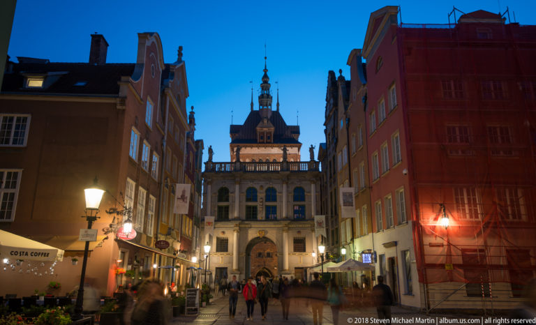Night view down Długa Street toward the illuminated Golden Gate between narrow historic façades and pedestrians.