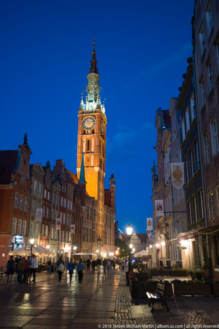 Illuminated Main Town Hall tower rising above gabled houses, pedestrians, and evening lights along Długi Targ.