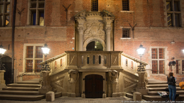 Symmetrical stone staircase with carved balustrades, sculpted supports, balcony, and ornate portal on a red-brick façade.