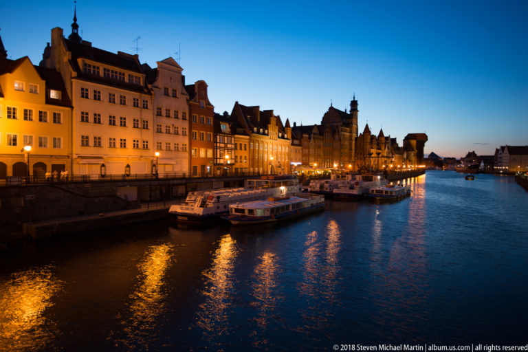 Illuminated Gdańsk waterfront with moored passenger boats, historic gabled façades, and golden reflections on the Motława River.