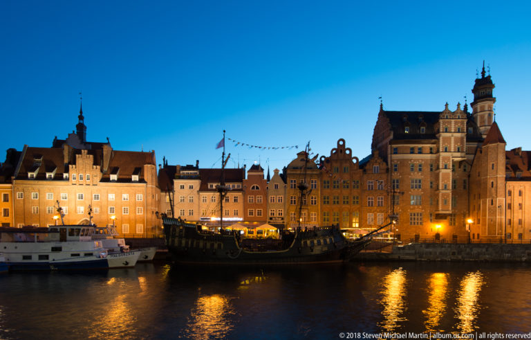 Dark historic-style sailing ship with masts and rigging moored before illuminated gabled buildings on the Motława waterfront.