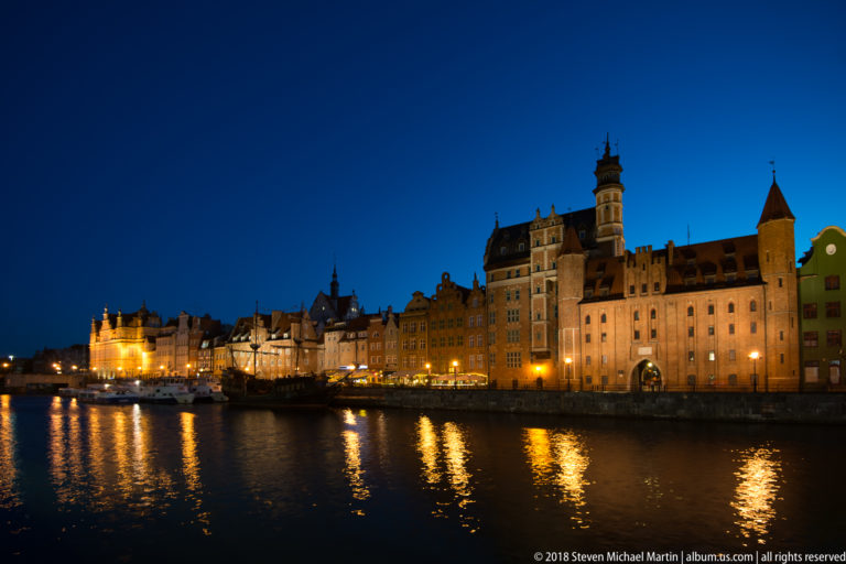 Nighttime Motława waterfront with illuminated historic façades, moored boats, and long golden reflections across the river.