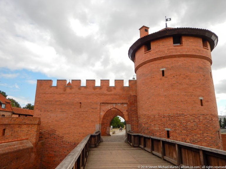 Wooden bridge leading to an arched brick gateway beside a large round defensive tower with narrow openings.