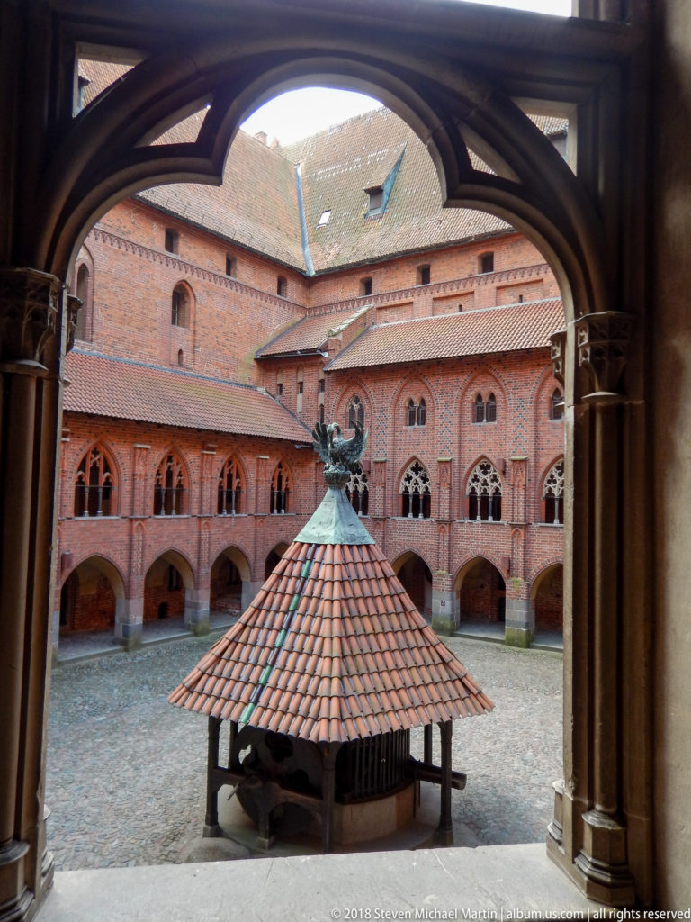 Courtyard and covered well seen through a dark pointed arch, with red-brick arcades and Gothic windows beyond.