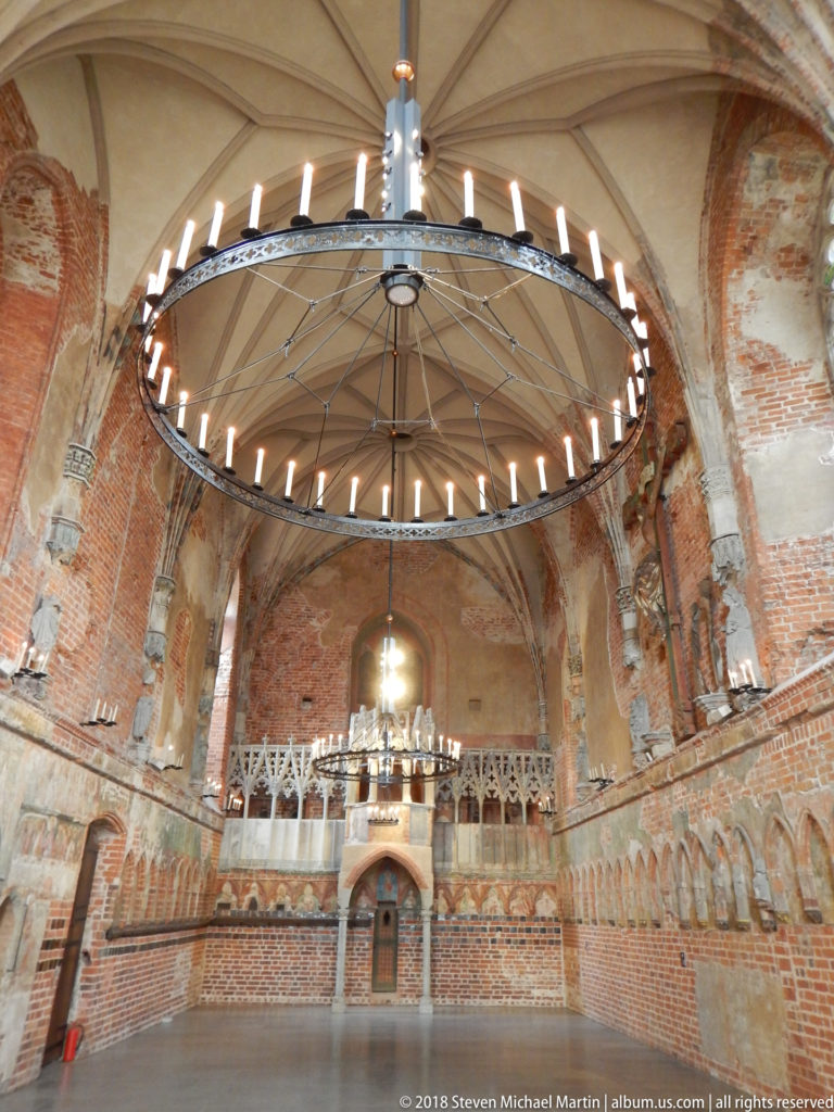 Large brick hall with pale rib vaults, a circular iron chandelier, and a raised gallery along the far wall.