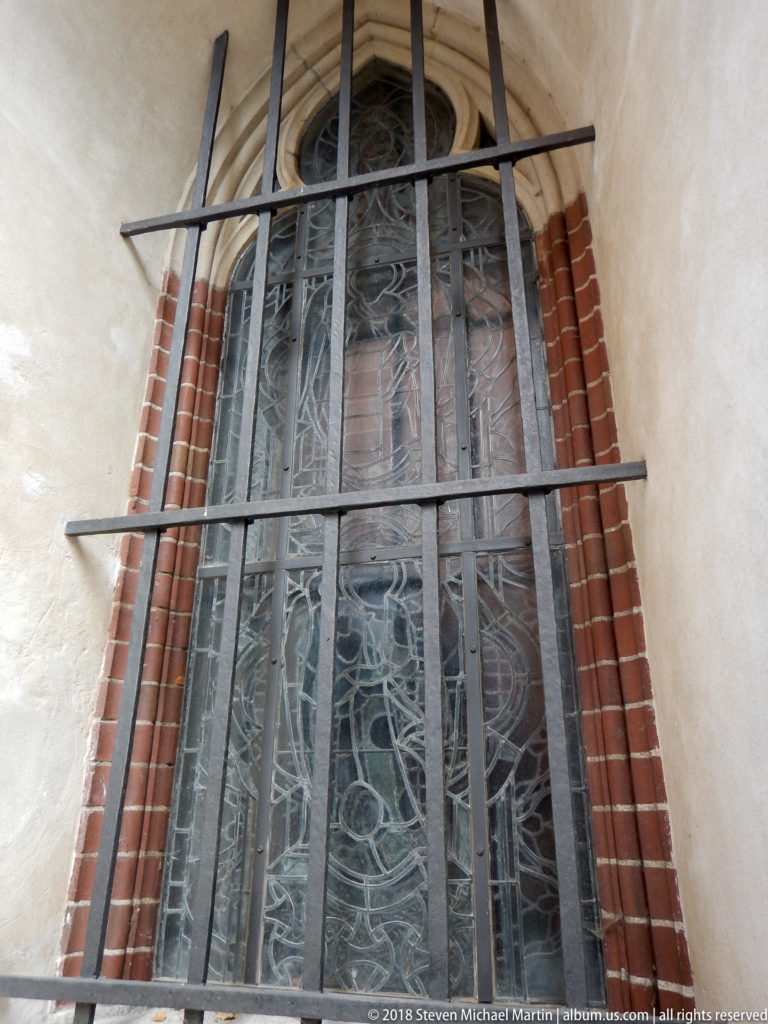 Tall pointed leaded-glass window in a brick reveal, crossed by dark protective bars and surrounded by pale plaster.