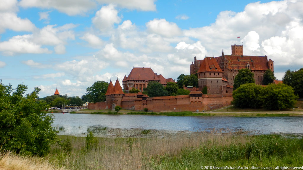 Malbork Castle across the Nogat River, with reeds in the foreground and red-brick walls, towers, and roofs beyond.