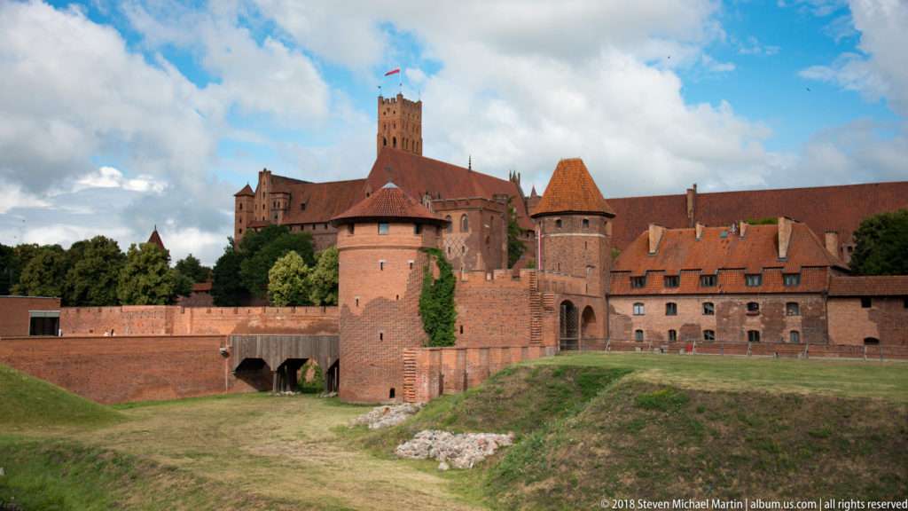 Round brick towers and outer walls in front of the taller castle core, viewed across a broad grassy ditch.