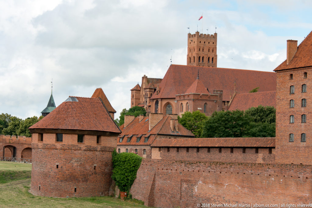 Low brick defensive tower and wall in the foreground with steep-roofed castle buildings and a tall square tower rising behind.