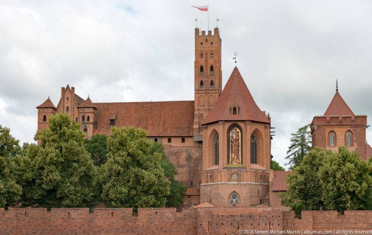 Tall Gothic brick projection with pointed windows below the castle’s square tower, partly screened by trees and an outer wall.
