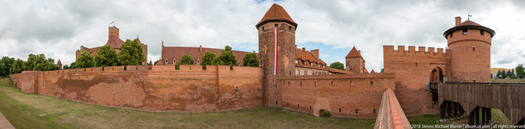 Wide view of Malbork Castle outer walls and towers, with a grassy ditch and bridge leading toward a round gate tower.