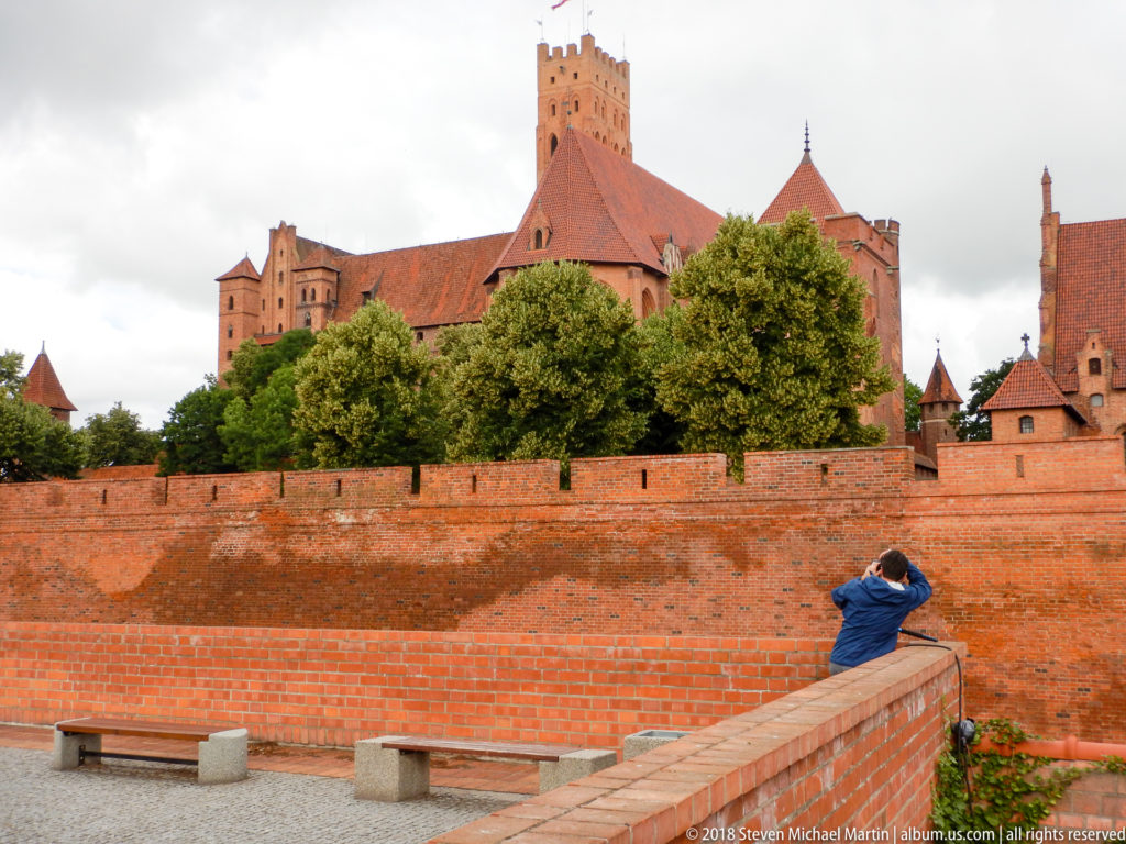 Layered red-brick castle buildings and towers rising behind an outer wall and trees, with a visitor in the foreground.