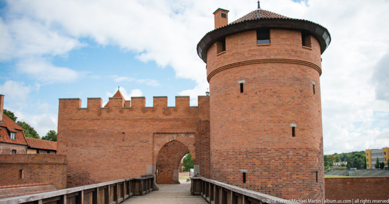 Wooden bridge leading to an arched brick gateway beside a massive round tower and crenellated wall.
