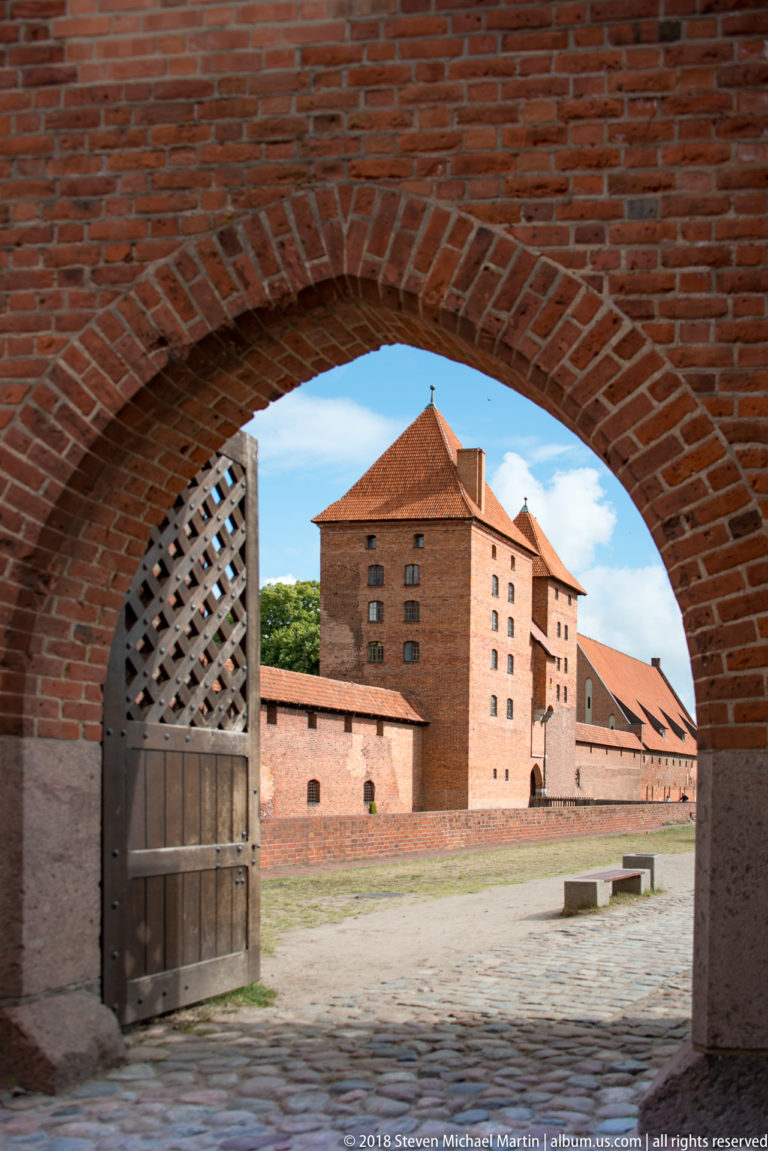 Sunlit castle courtyard and brick buildings seen through a shaded brick gateway with a heavy timber door.