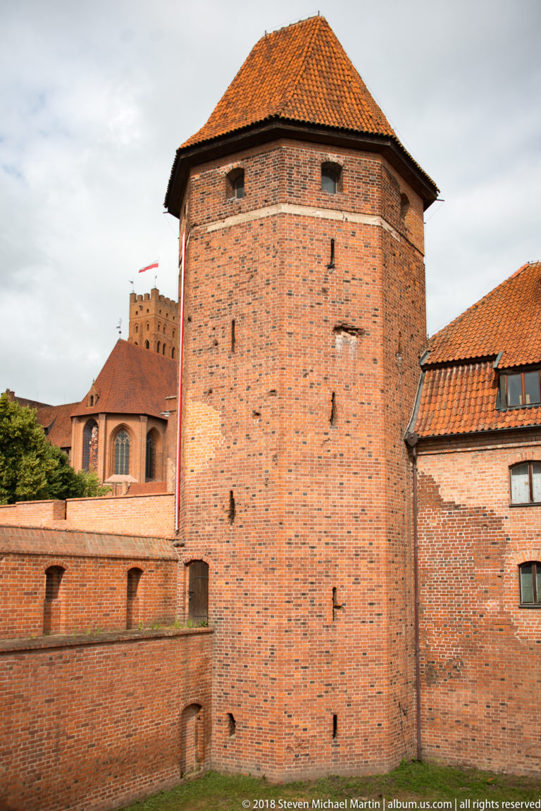 Large polygonal red-brick defensive tower with a hipped tile roof, joined to lower walls with castle buildings behind.