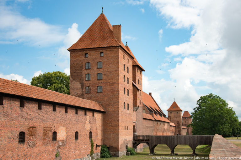 Tall rectangular red-brick tower above lower castle ranges, with small windows, tiled roofs, and a timber bridge-like connector.