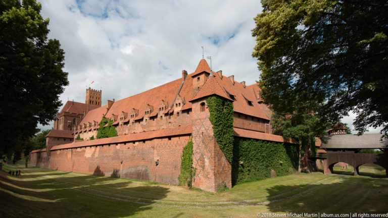 Long red-brick fortification wall with a large angled buttress and ivy, beneath steep castle roofs and a distant tower.