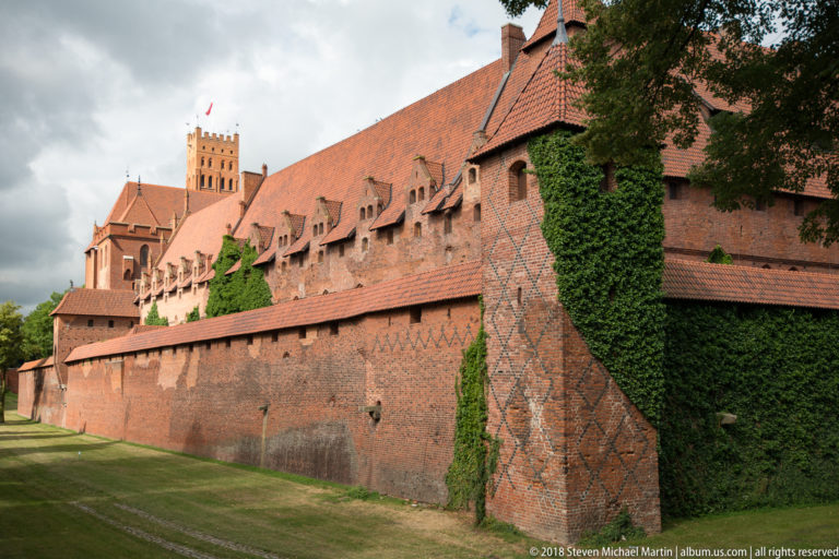 Large patterned brick buttress and ivy-covered outer wall below a steep-roofed castle range with small dormers.