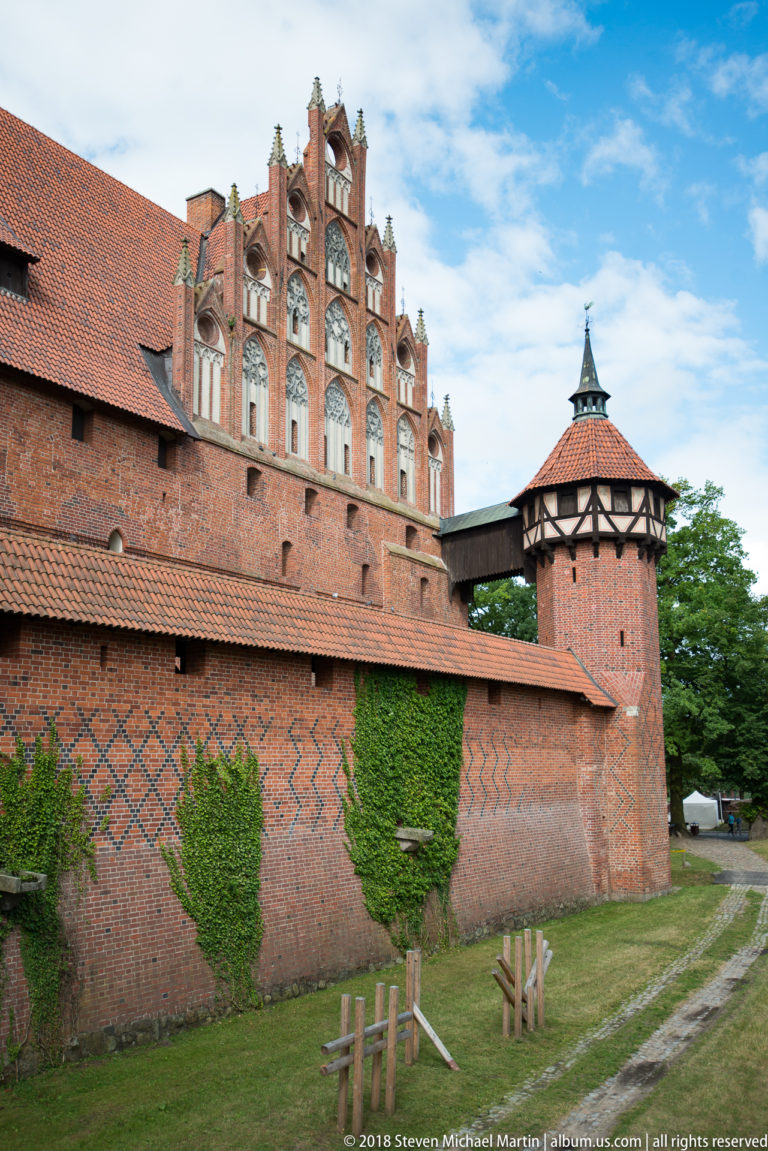 Ornate Gothic brick gable above a patterned outer wall beside a small timber-framed corner tower with a pyramidal roof.