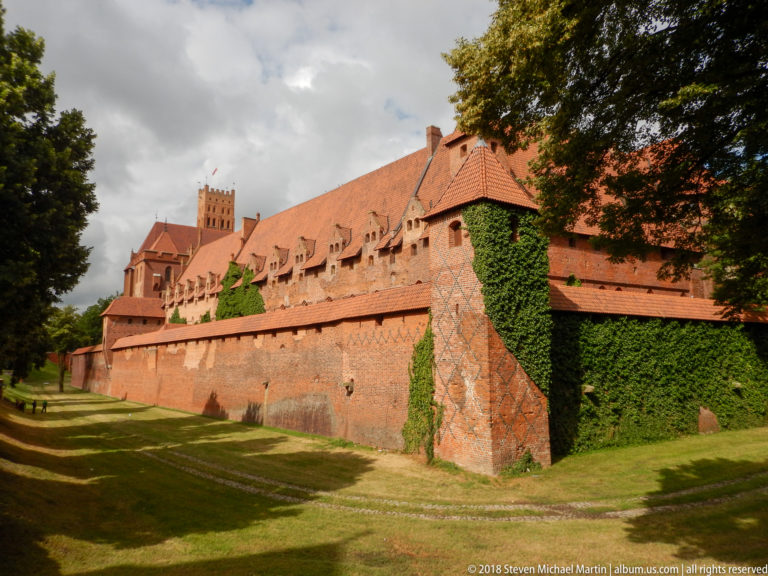 Long red-brick defensive wall below a steep-roofed castle wing, with buttress, ivy, dormers, and grass in front.