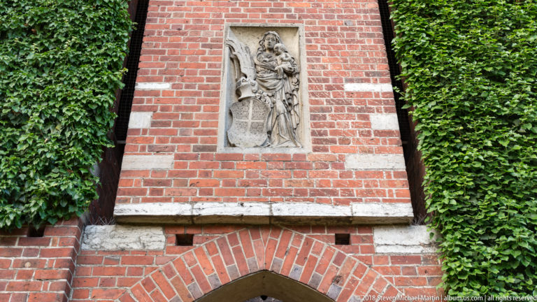 Carved stone religious relief set into a red-brick wall above a pointed arch, with thick ivy climbing on both sides.