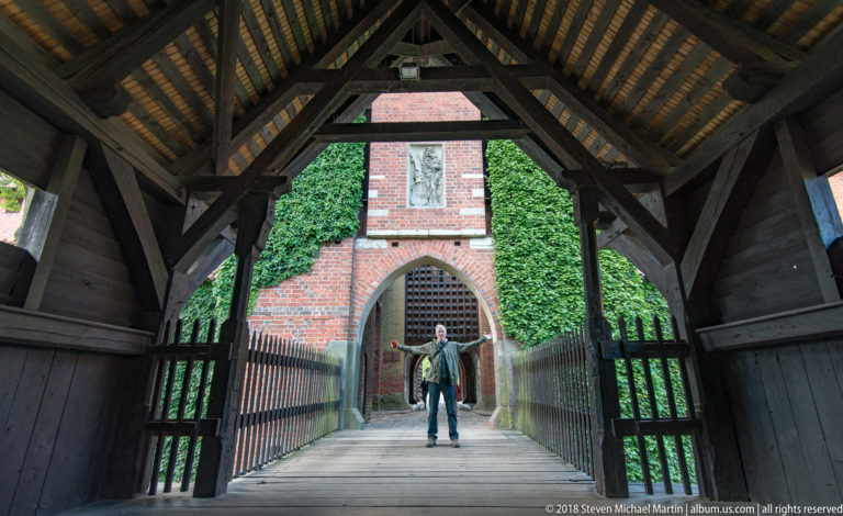 Timber-covered passage with exposed roof trusses leading toward a pointed brick gateway, where a visitor stands near the entrance.