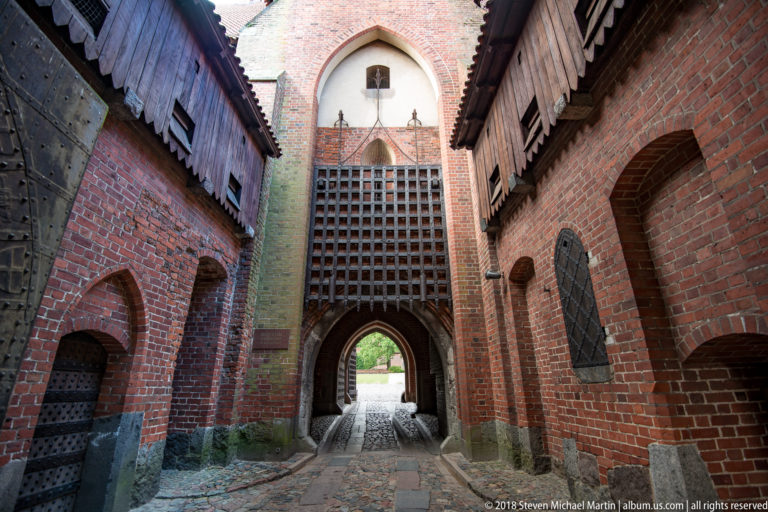 Narrow brick gateway interior with a large timber grille, pointed arches, heavy doors, and a bright opening at the far end.