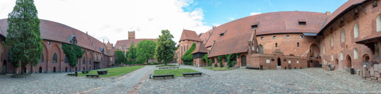 Wide cobbled courtyard enclosed by red-brick castle ranges with steep tiled roofs, towers, trees, benches, and grass.