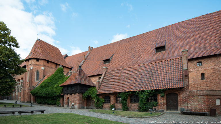 Long red-brick castle range with steep tiled roof, projecting structures, a polygonal tower, and ivy along the lower walls.
