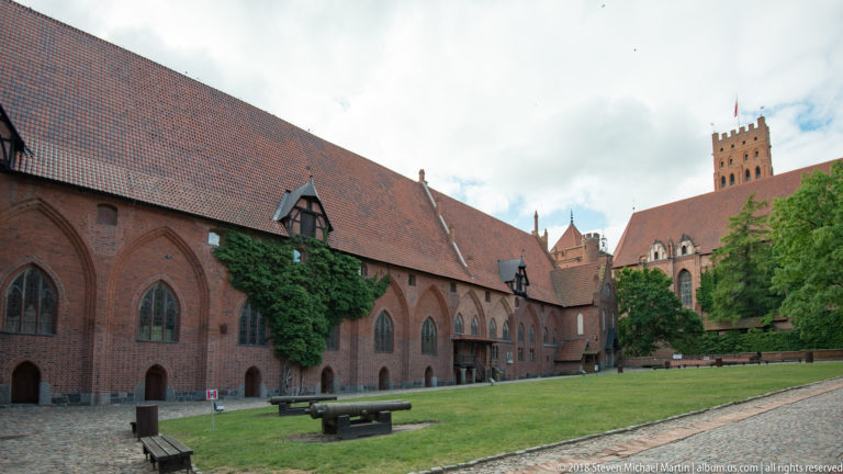 Courtyard-side red-brick range with tall pointed windows, ivy, steep tiled roof, benches, grass, and towers beyond.