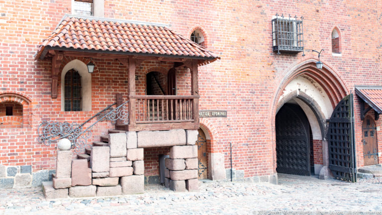 Pointed brick doorway beside a small roofed wooden balcony, with stacked stone blocks and barred windows in the courtyard wall.