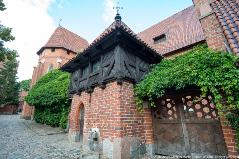 Small brick and half-timbered roofed structure with an open arch and wooden gate, set against ivy-covered castle walls.