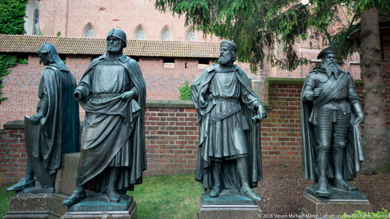 Four life-size dark bronze statues of historical figures standing together before a red-brick castle wall and trees.