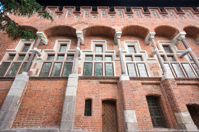 Upward view of a red-brick Gothic façade with pale stone-framed windows, recessed arches, and blind arcading below the roofline.
