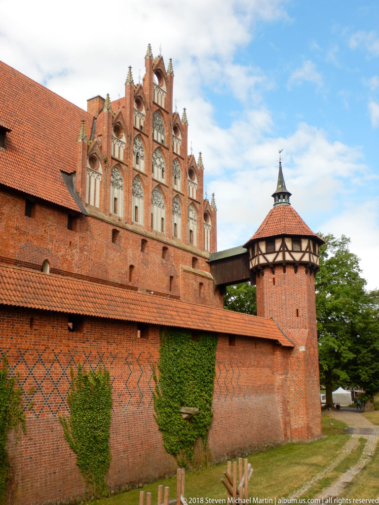 Ornate Gothic brick gable above a defensive wall beside a small timber-framed tower with a pyramidal roof.