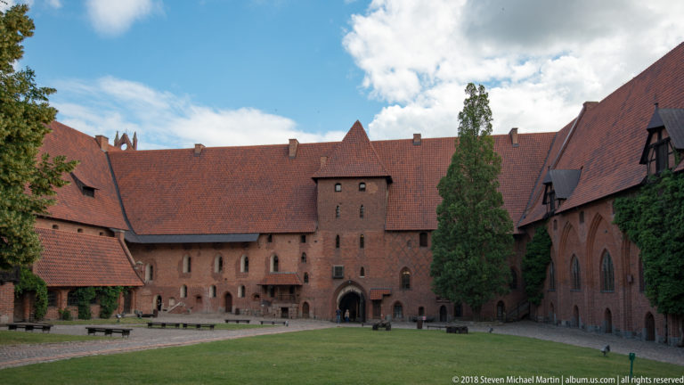 Large grassy courtyard enclosed by red-brick castle ranges, with steep tiled roofs, a central arched entrance, and tall trees.